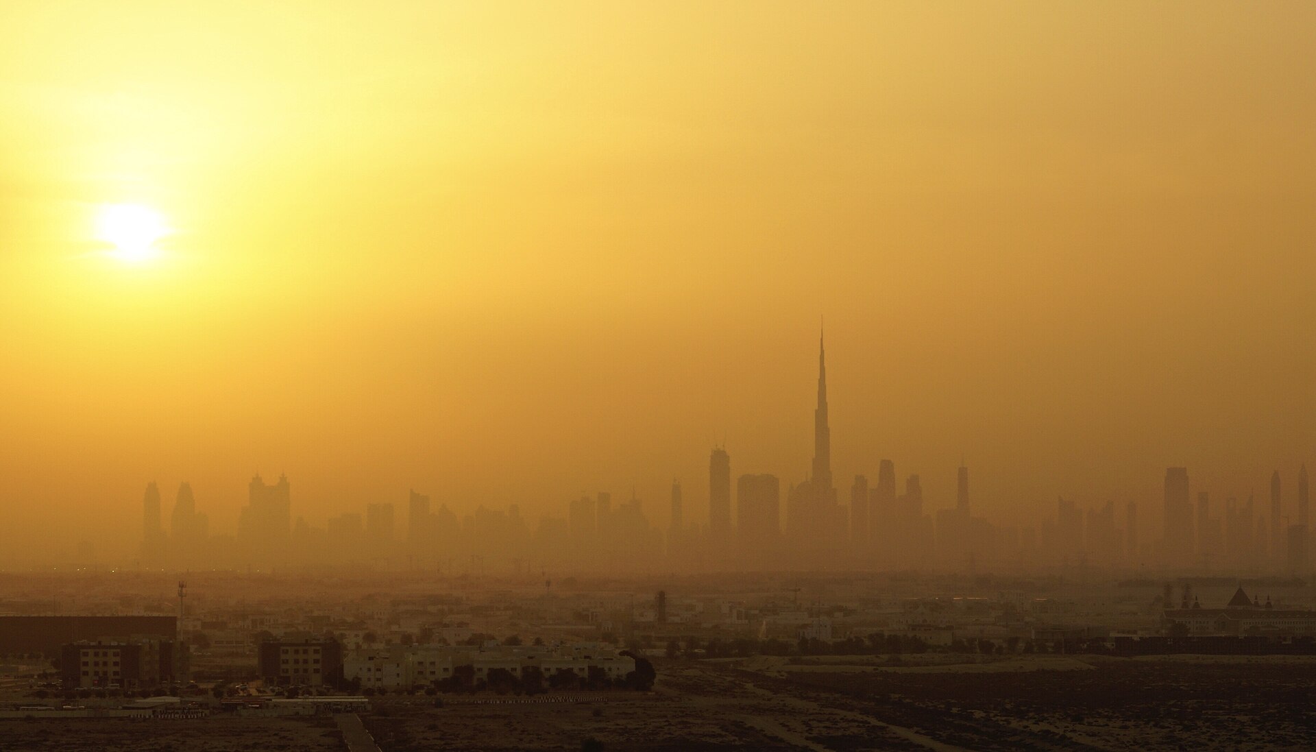 The Dubai skyline in a gold haze at sunset, seen from the desert edge of the city.